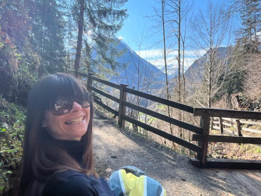 A person sitting on a bench smiling, with a fence, mountains and blue skies in the background.