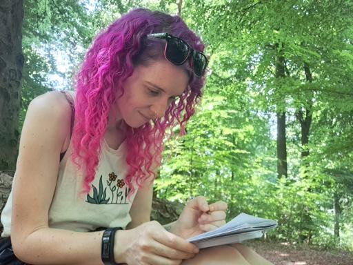 A female with pink curly hair and sunglasses on her head, sitting in a woodland. She is writing in a notepad and smiling.
