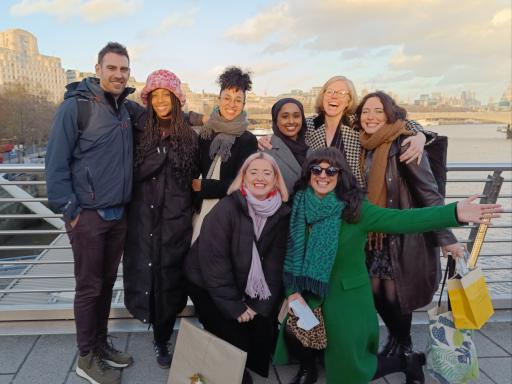 A group of eight people in winter clothing standing on a bridge looking to camera. They are smiling and laughing with arms around each other