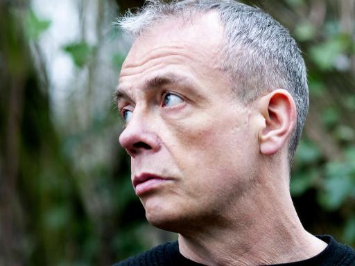 A white male which a closely shaved head looking to his right amongst a tree blurred background