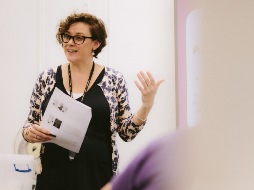 A white woman with short brown curly hair, wearing glasses and black dungarees presenting to a class