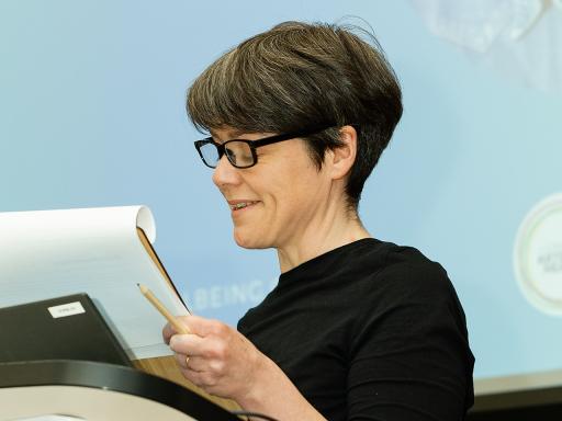 a woman with short brown hair and glasses reads at a podium