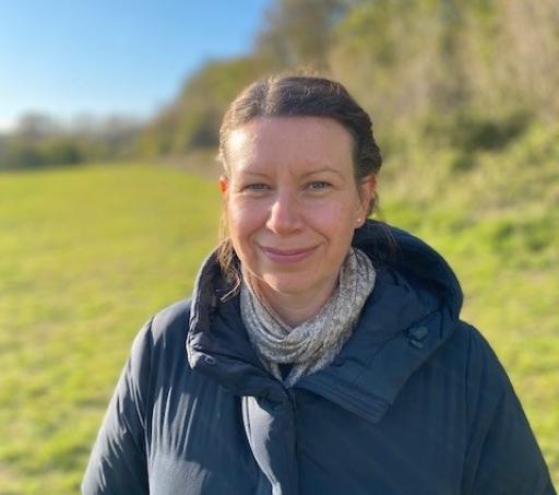 A white woman wearing a puffer jacket smiling at the camera with her hair tied back standing in front of a green field