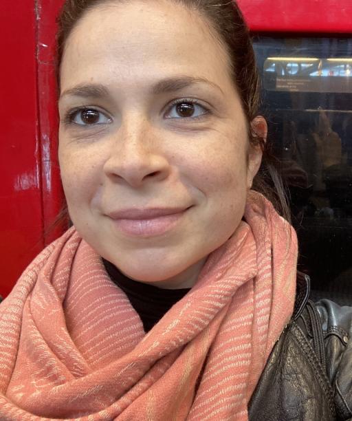 A woman smiling facing the camera wearing a salmon pink scarf with her hair tied back, leaning against a red door frame