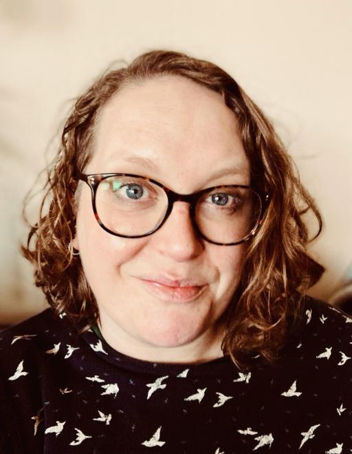 A white woman with short brown curly hair, wearing glasses and black dungarees presenting to a class