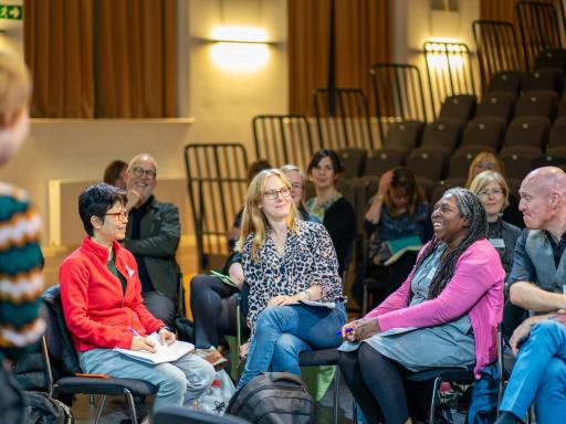 A white woman wearing glasses with short black hair and a red top sitting next to a white woman with shoulder length blonde hair who is sitting next to a black woman with locs wearing a pink cardigan smiling