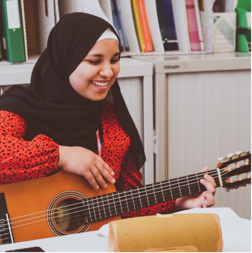A young muslim woman smiling wearing a black hijab and a red top playing a brown guitar