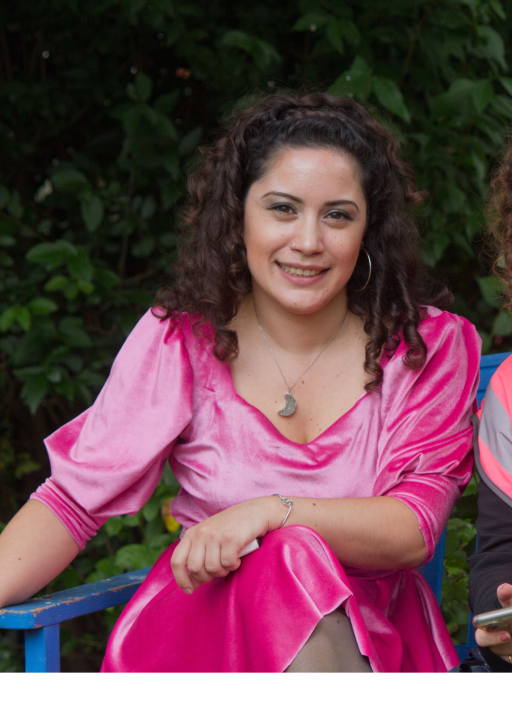 An olive skinned woman with dark, curly, brown hair wearing a bright fushia dress sitting on a blue chair outside smiling