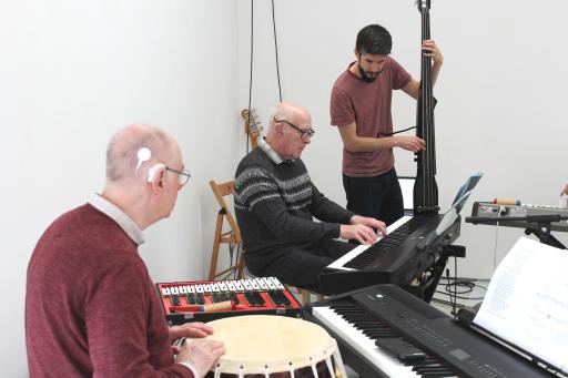 Two older white men sitting down playing the drums and the keyboard, younger bearded male standing playing an instrument