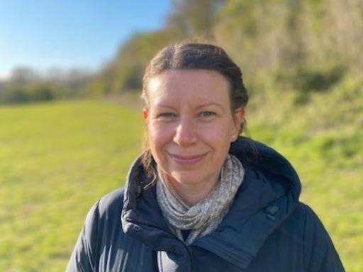 A white woman wearing a puffer jacket smiling at the camera with her hair tied back standing in front of a green field