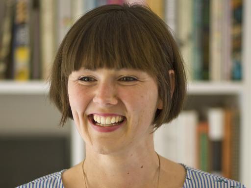 A white woman with a short brown bob wearing a blue dress smiling  in front of a bookcase