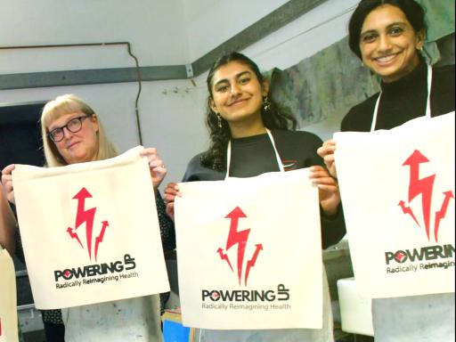 Five people smiling to camera, holding white totebags with a red logo and 'Powering Up' written in black