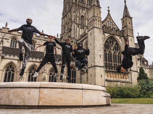 Four black in black sports clothes teenagers jumping midair in front of Gloucester Cathedral