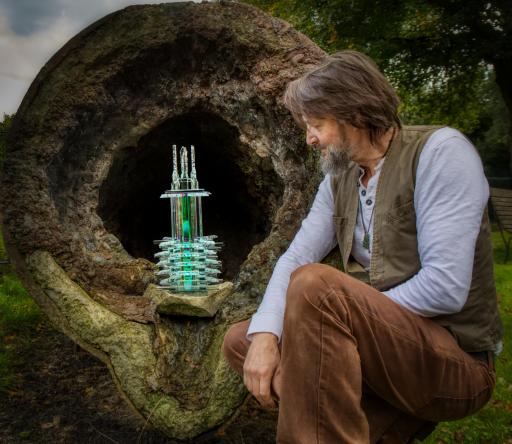 A man with long brown hair wearing brown trousers and a light blue shirt looking at a glass sculpture resting within a tree outside in nature