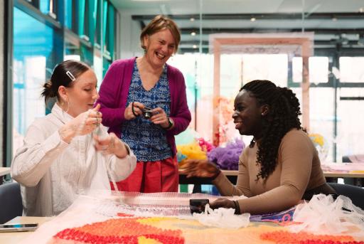 two women sitting down at a table smiling at each other doing arts and crafts with a woman standing between them smiling