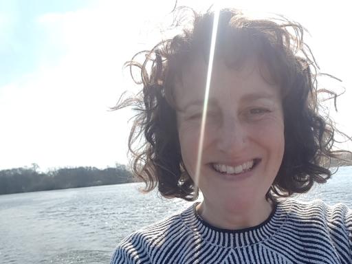 a woman wearing a stripey jumper with curly brown hair smiling at the camera against a sea background
