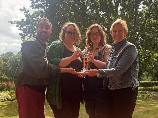 Four people, the leadership team of Next Door But One, standing in the sun in front of trees looking happy, all holding a bright and colourful glass award with ‘CHWA -  Culture Health and Wellbeing Alliance’ on it