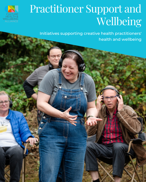 A white woman wearing navy dungarees over a grey t-shirt is wearing headphones and smiling in a woodland area with multiple people sitting in the background