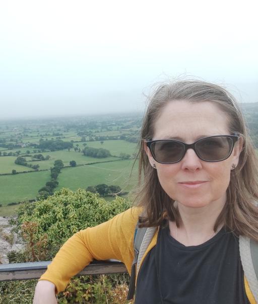 A white woman with shoulder length brown hair wearing sunglasses, a black top and yellow cardigan facing the camera and smiling, as she leans against a railing with amidst a natural green background