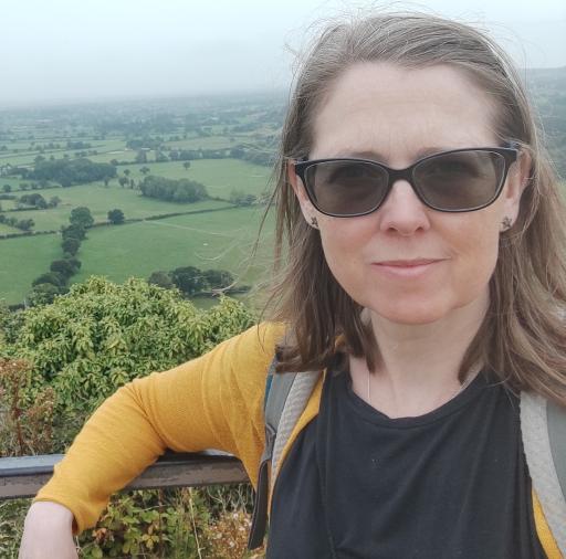A white woman with shoulder length brown hair wearing sunglasses, a black top and yellow cardigan facing the camera and smiling, as she leans against a railing with amidst a natural green background