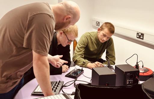 Two young white males sitting down at a desk with an older white man standing over them whilst they engage with music equipment