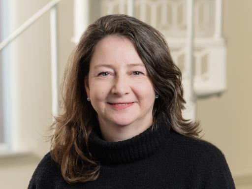 Half-length portrait photo of a woman with long dark hair and a black jumper stannding in front of white bannisters