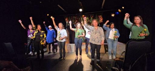 A group of people wearing multi-coloured clothing standing on a stage with dim lighting standing with their hands in the air