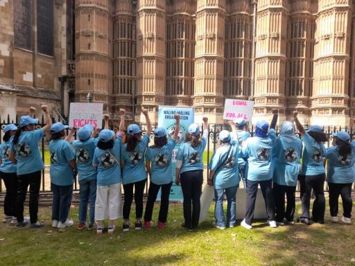 a group of people in blue t-shirts stand with fists raised in front of the houses of parliament carrying placards 