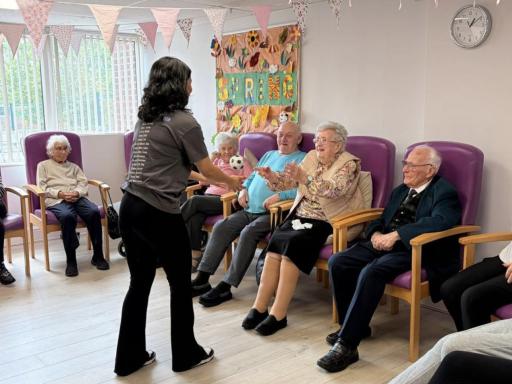 A woman with long black hair wearing a grey t-shirt and black trousers standing and gesturing to older people sitting in purple chairs facing her