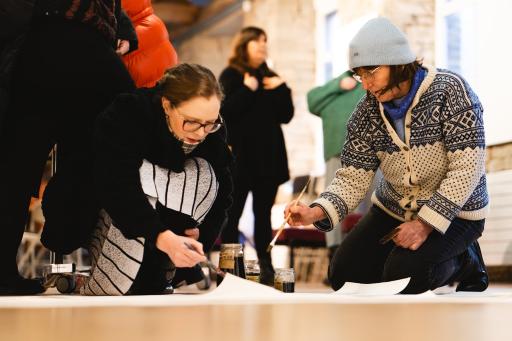 Two women wearing glasses and thick patterned wooly jumpers kneeling down to paint on a large sheet of paper