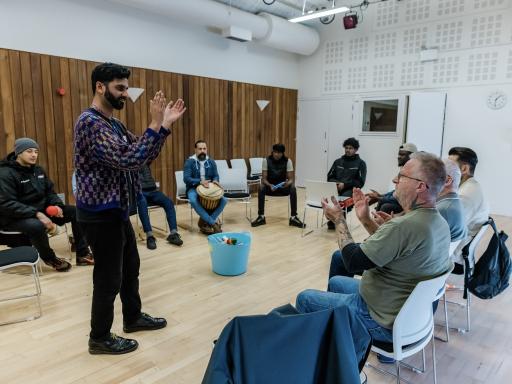 A bearded male with short black hair wearing a dark coloured wooly jumper with flecks of colour and black jeans standing in front of seated individuals in a large community space with his hands held in a clapping stance