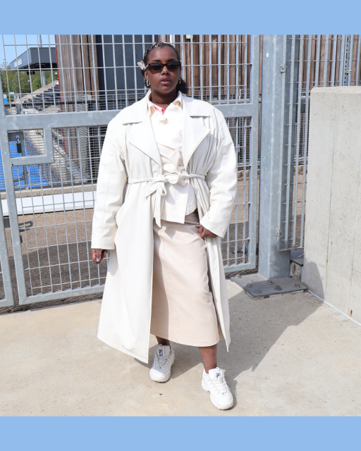 A black woman wearing a white jacket, black sunglasses, a white skirt and trainers posing, facing the camera
