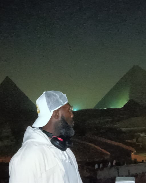 A black man wearing a white baseball cap backwards and black headphones facing away from the camera into the distance against the backdrop of egyptian pyramids