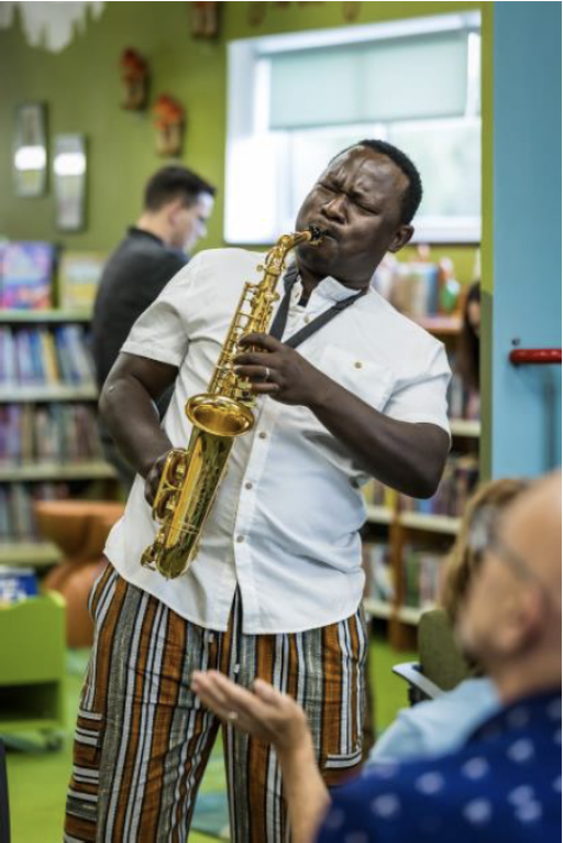 A black man wearing a white shirt and striped trousers playing a saxophone in a library environment