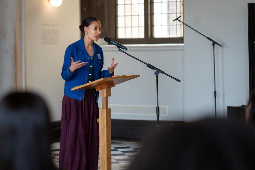 A woman standing at a lecturn speaking into a microphone wearing a royal blue shirt and a purple skirt